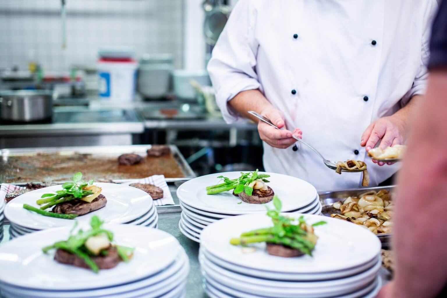 Stacked plates with nicely-presented meat on top, with a chef holding a spoon plating vegetables