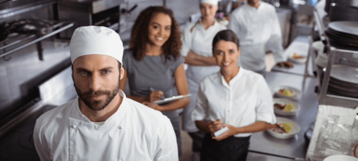 A male chef headshot in a kitchen with two women holding clipboards behind him.