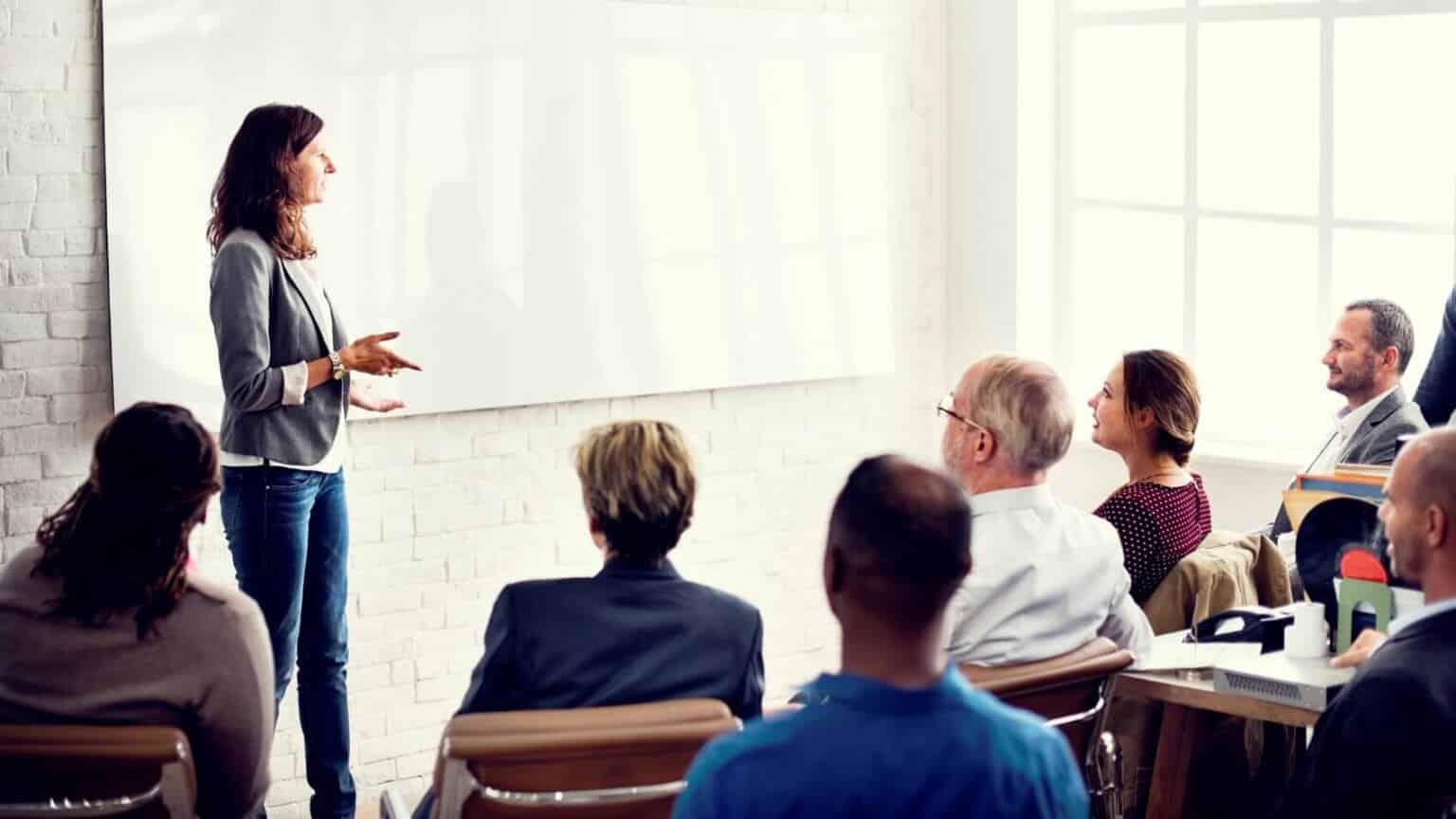 Woman presenting in front of a whiteboard to 7 people sitting in chairs