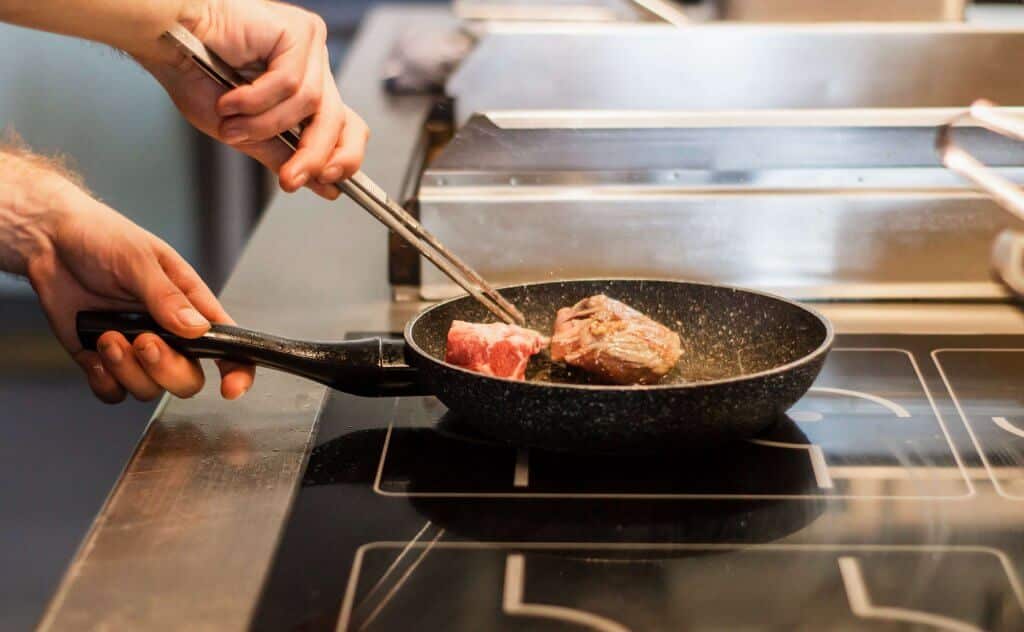 Hands holding metal utensil to flip meat in a hot pan