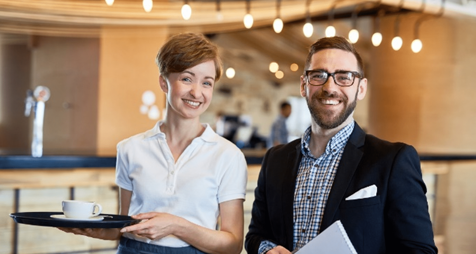 A woman with short hair holding a tray with coffee and a man with glasses holding papers, both smiling